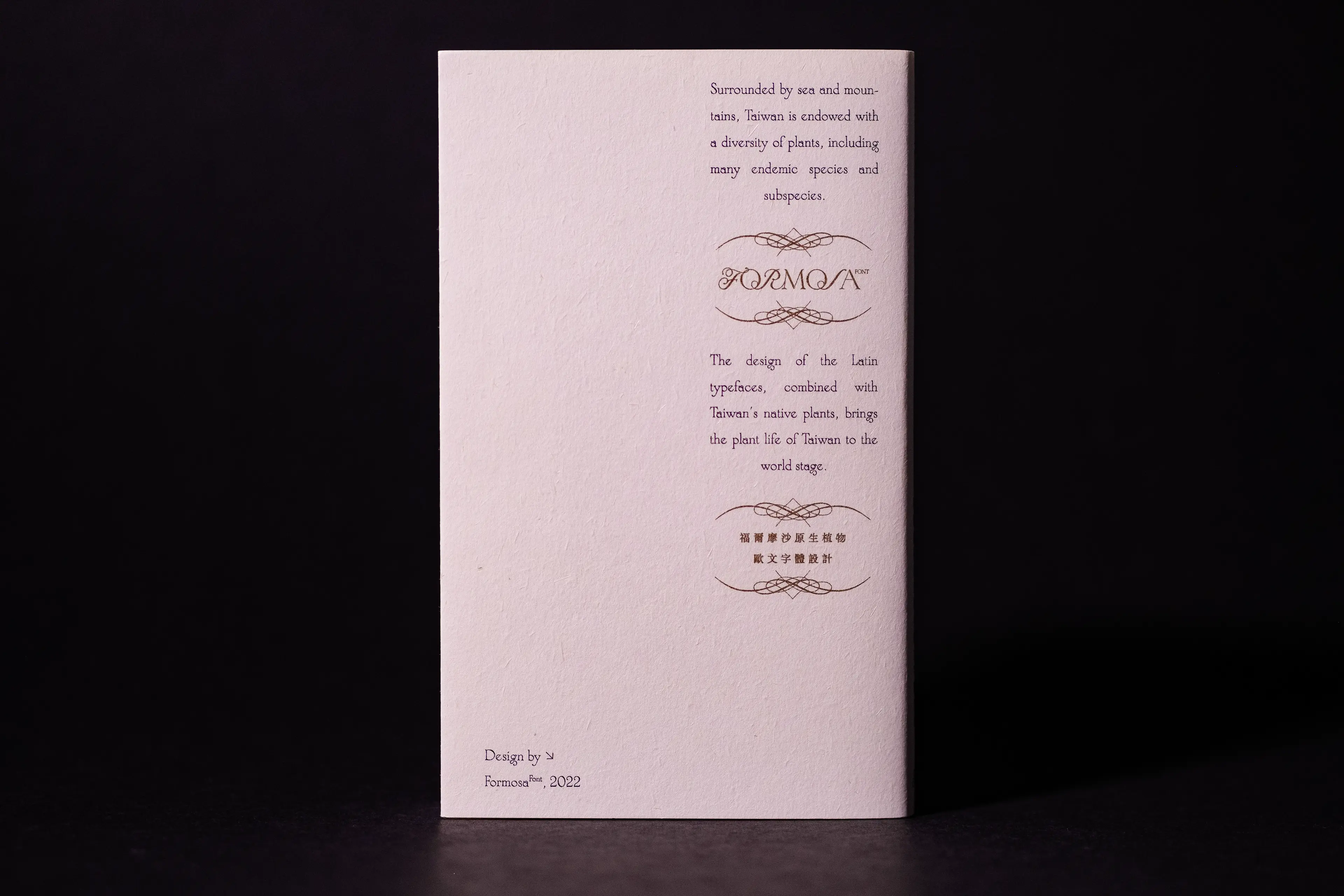 The back cover of a slim white booklet photographed against a dark background, featuring the gold-foil "FORMOSA" logotype flanked by ornate flourishes, with bilingual body text in Latin and Chinese describing Taiwan's endemic plant life and the typeface's design concept.