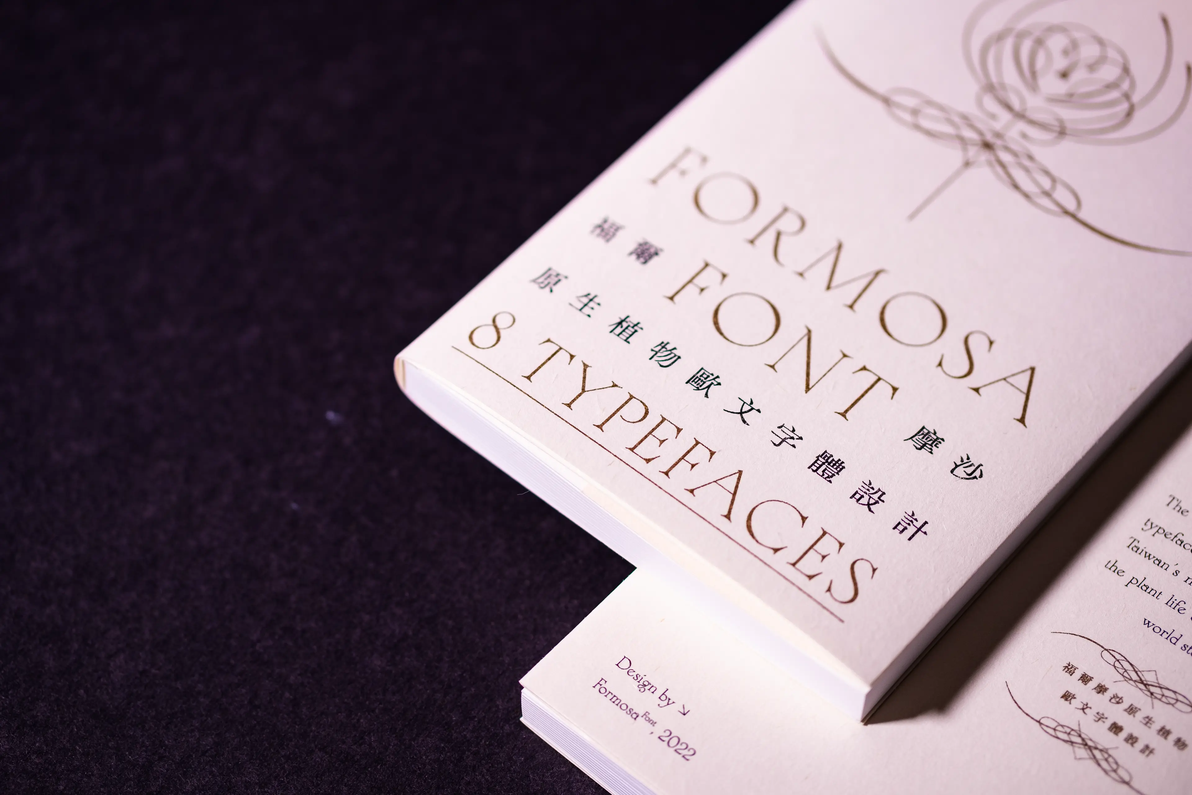 A printed booklet titled "Formosa Font — 8 Typefaces" lies at an angle on a dark surface, its cream-white cover featuring elegant serif Latin lettering alongside vertical Chinese text, with a delicate botanical line illustration in the upper right corner.