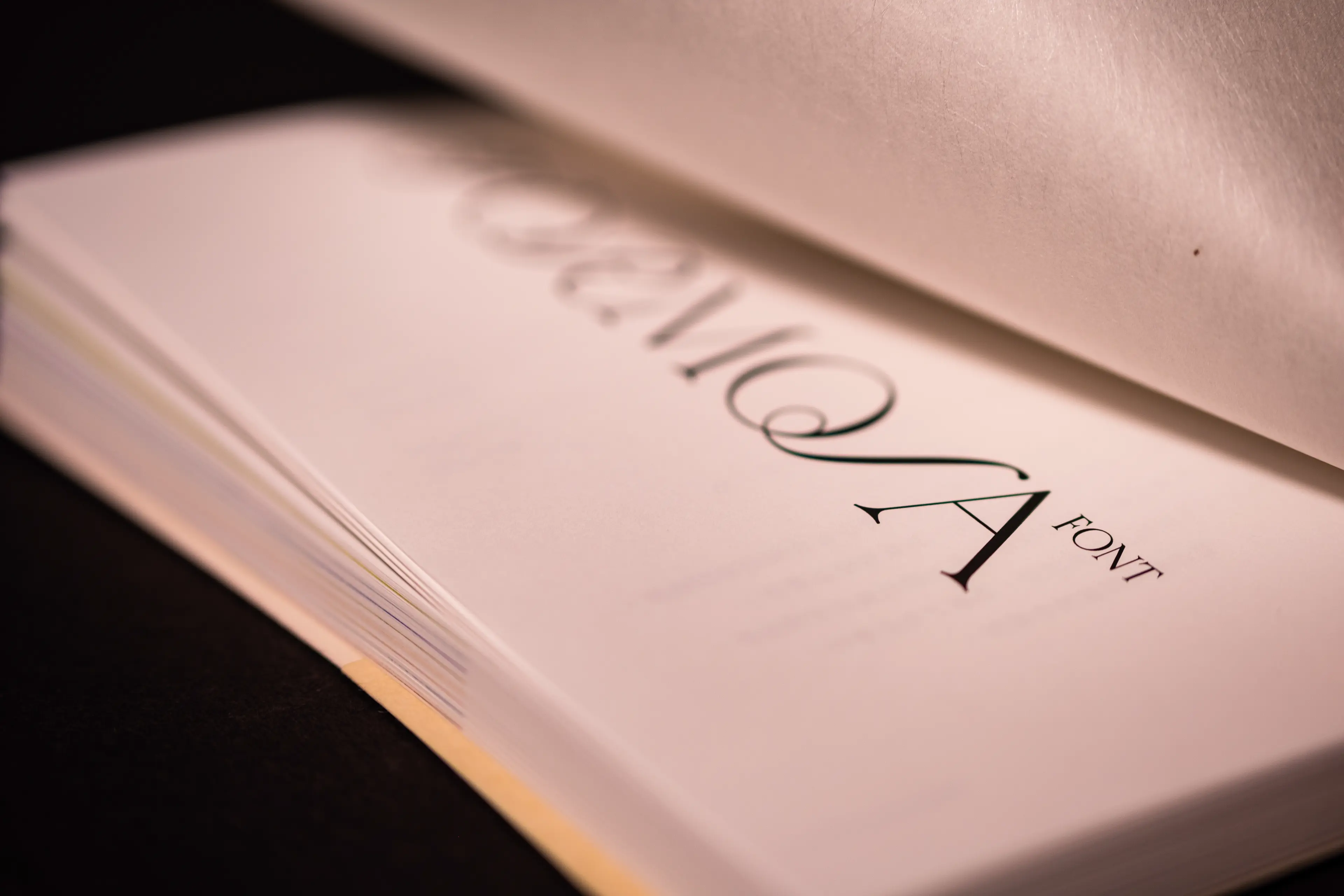 A close-up photograph of a thick white-paged book opened to reveal its cover, displaying an elegant serif typeface with a stylized "A" and the word "FONT" in refined letterforms. The shallow depth of field and dark background emphasize the crisp, high-contrast black typography against the bright white paper.