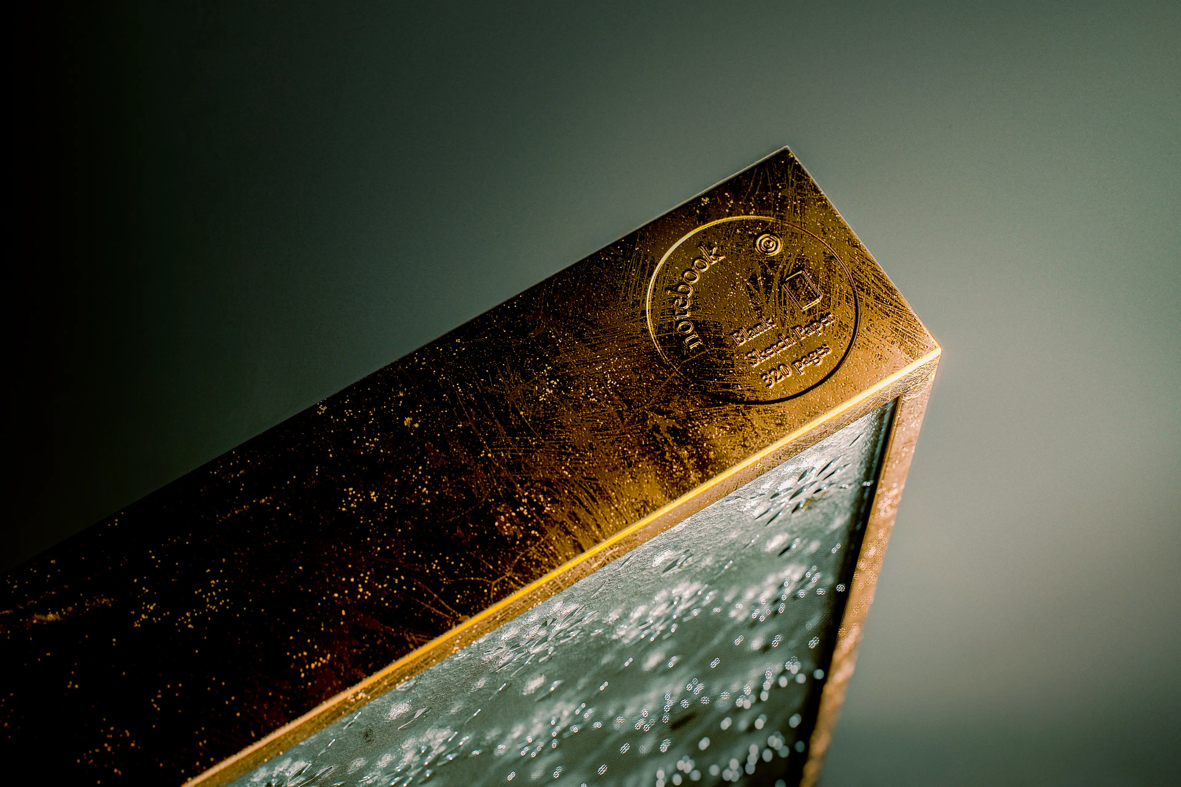 Close-up of the notebook's top corner, highlighting a weathered brass-toned metal spine with an embossed circular logo, set against a textured glass cover with a raindrop-like bubble pattern.