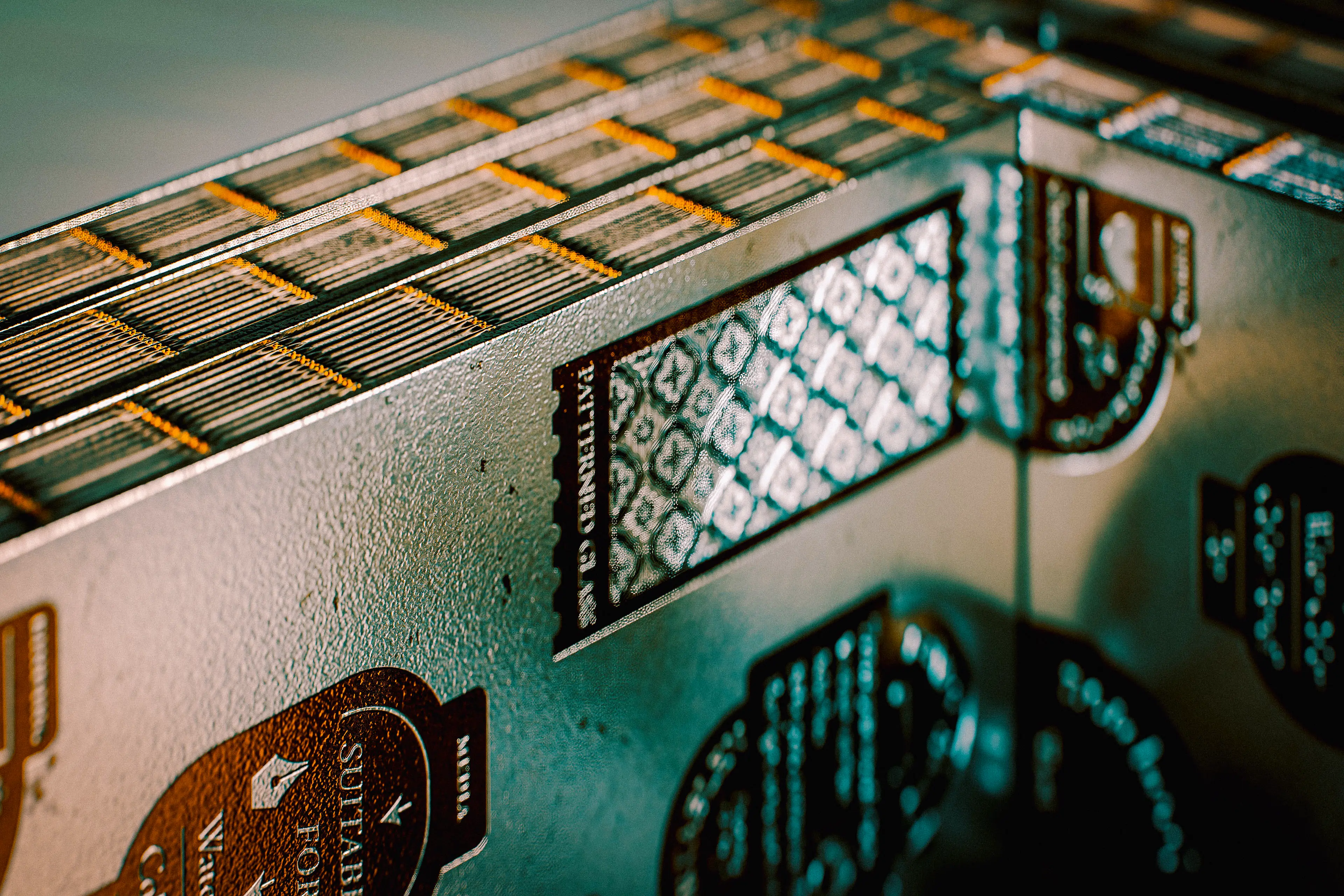 A close-up of the packaging box's corner, revealing layered notebook spines with gold-accented edges stacked inside, while the box exterior displays an intricate geometric tile pattern in dark teal and black with embossed badge-style typography.