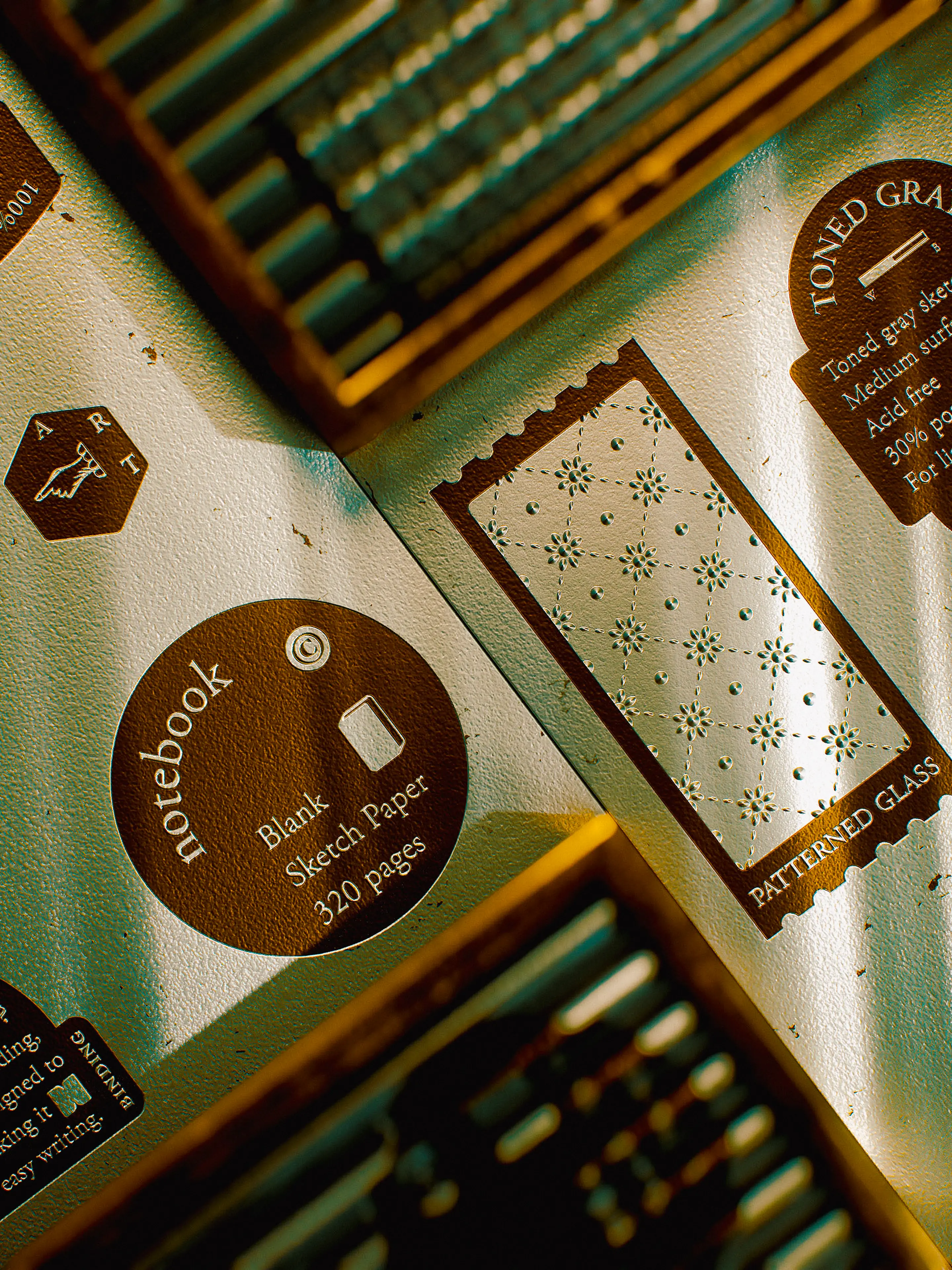 Close-up of the notebook's back cover details under warm, raking light, showing a circular brown label reading "notebook — Blank Sketch Paper — 320 pages" alongside a stamp-shaped vignette of the patterned glass motif and a "PATTERNED GLASS" text badge, all printed in white on a warm brown ground.