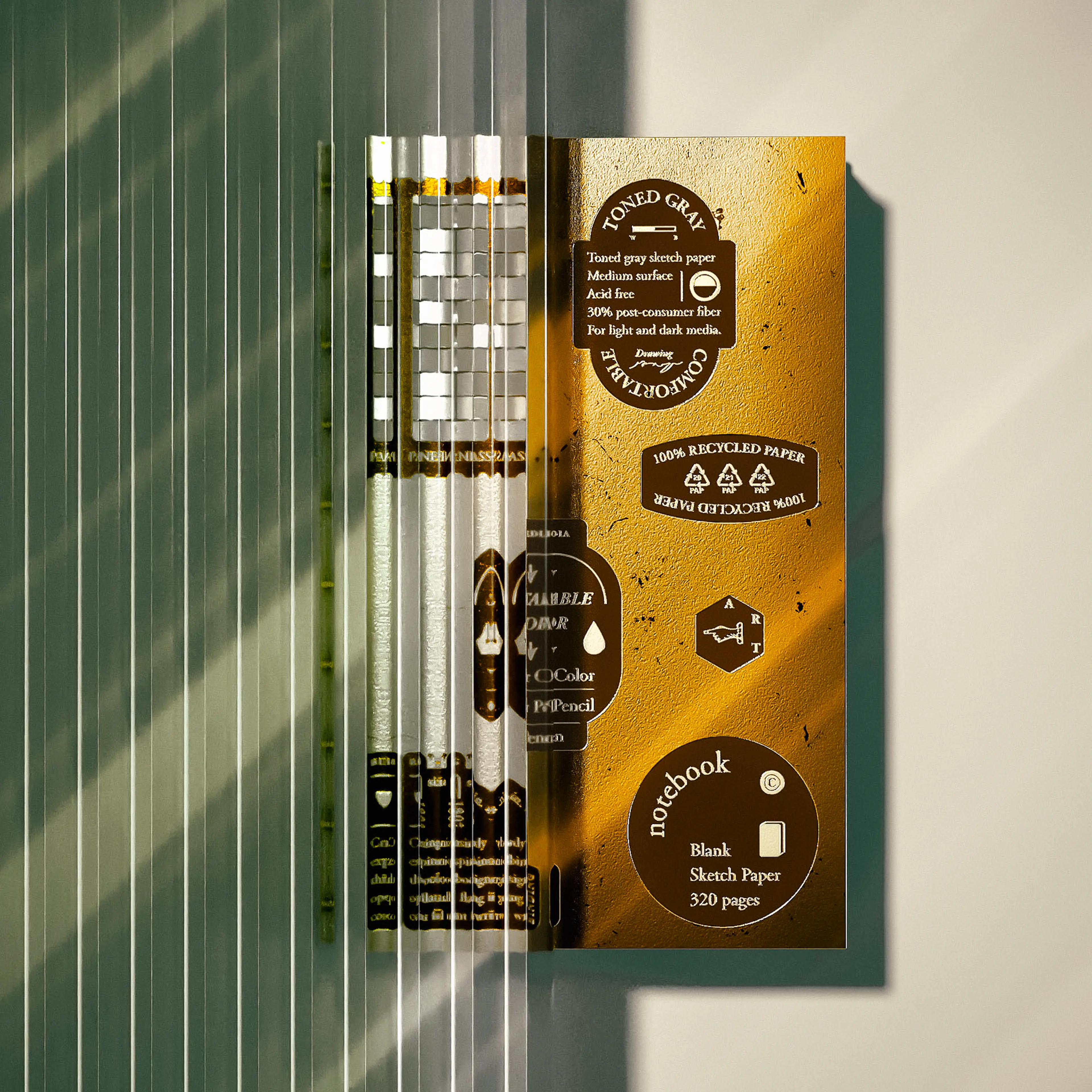 A gold-toned notebook back cover displaying product specification badges — including "Toned Gray," "100% Recycled Paper," and "Blank Sketch Paper 320 pages" — photographed through ribbed glass that refracts the image with vertical stripe distortions and soft shadow accents.